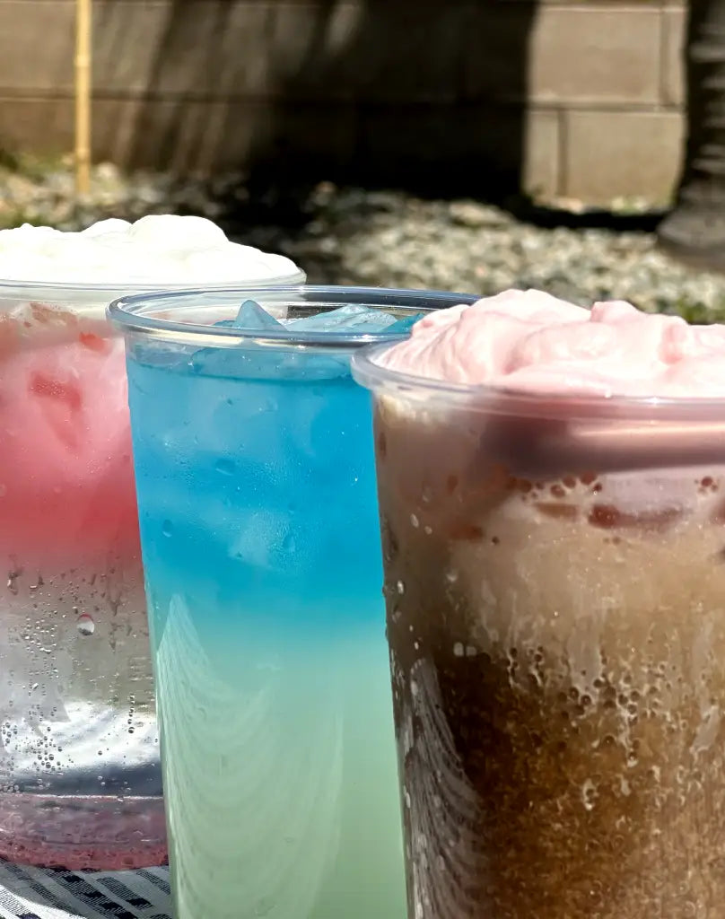 Three colorful drinks in clear cups on a table: a pink layered drink with whipped cream, a blue icy drink, and a brown fizzy drink topped with pink foam.
