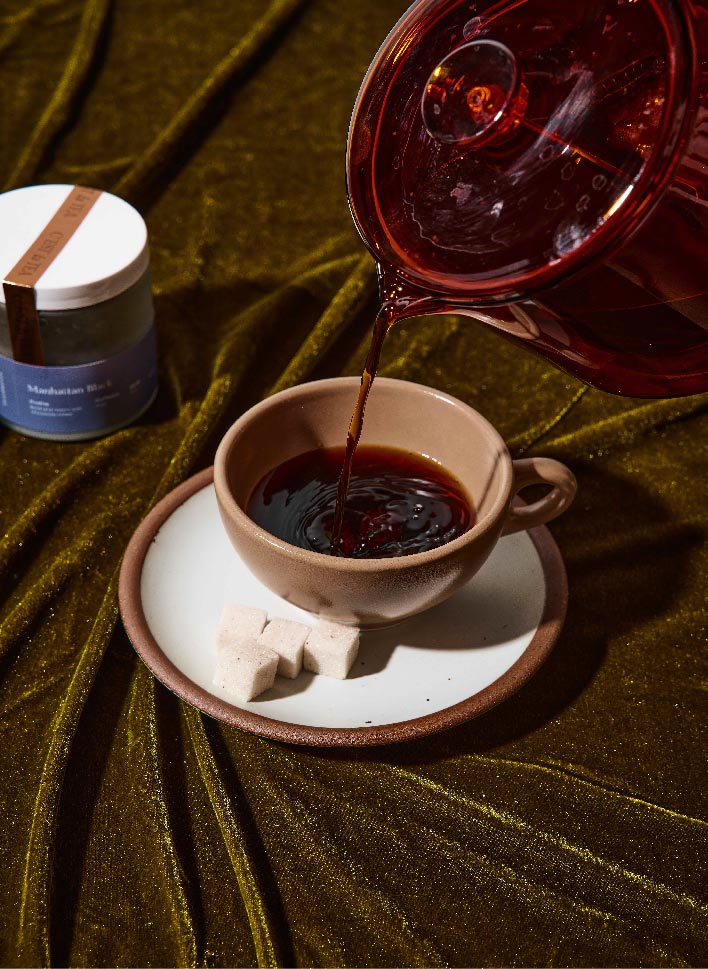 A cup of coffee is being poured from a glass pot, with sugar cubes on a plate beside it, set against a textured green fabric background.