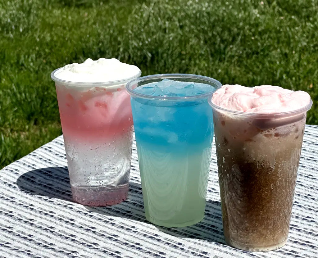 Three colorful drinks in plastic cups: a pink and white layered drink on the left, a blue drink in the center, and a brown drink topped with pink foam on the right, set against a grassy background.